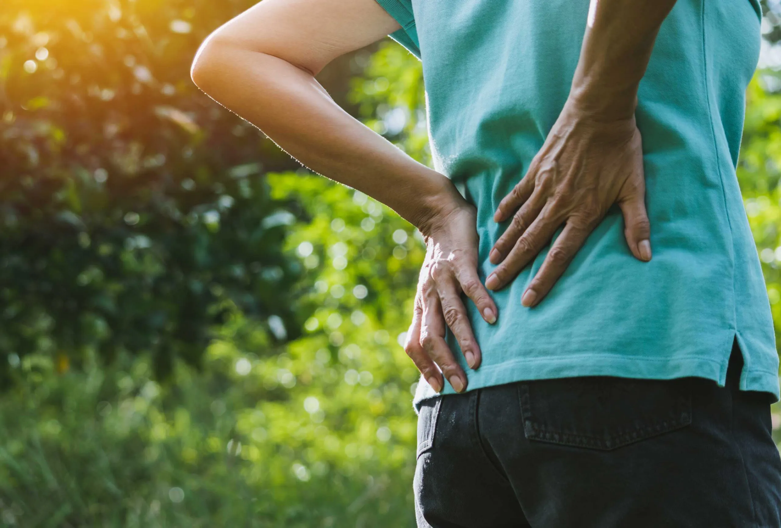 a woman in a teal shirt holding her back in pain outside in a park